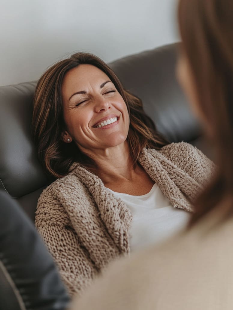 A woman with brown hair is leaning back on a dark sofa, smiling with her eyes closed. She is wearing a beige knit cardigan over a white top. Another person, slightly out of focus and with brown hair, appears in the foreground—perhaps they're discussing their experience at the Online Counselling Australia.