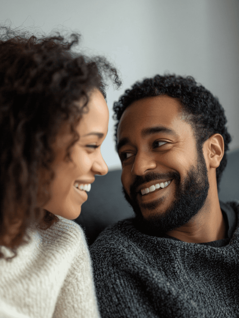 A couple is smiling at each other, sitting close together on a couch. The woman has curly hair and is wearing a light-colored sweater, while the man has short curly hair and a beard, and is wearing a dark sweater. The background is softly blurred, creating an intimate setting reminiscent of Online Counselling Australia sessions.