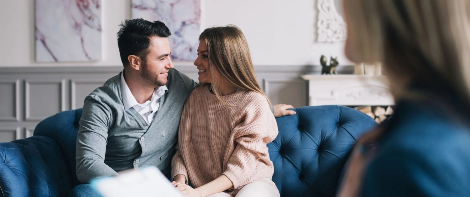 A couple sits closely together on a blue sofa, smiling and looking into each other's eyes. A third person, slightly out of focus and holding a clipboard, is seated in the foreground, possibly suggesting a therapy or counseling session. The room has elegant decor.