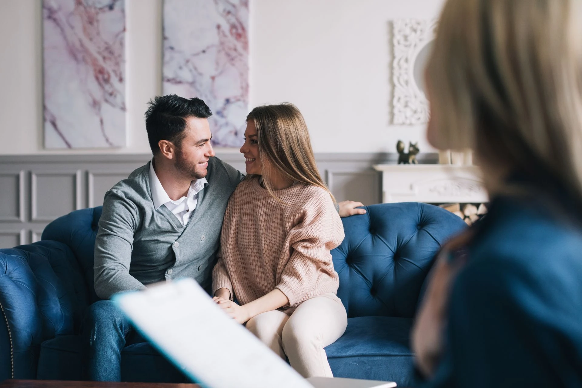 A couple sits closely together on a blue sofa, smiling and looking into each other's eyes. A third person, slightly out of focus and holding a clipboard, is seated in the foreground, possibly suggesting a therapy or counseling session. The room has elegant decor.